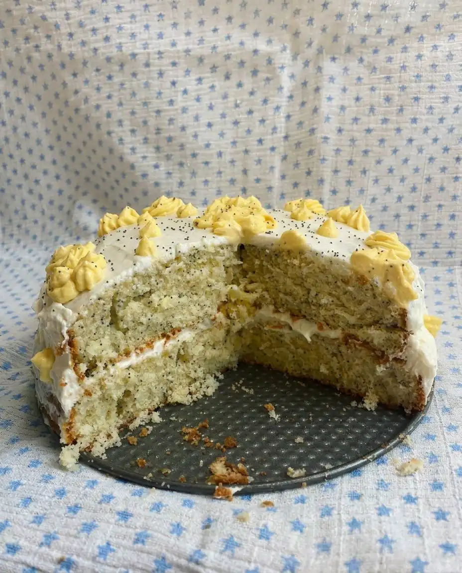 A hand using a spatula to spread thick white cream cheese frosting over a yellow poppy seed sheet cake.