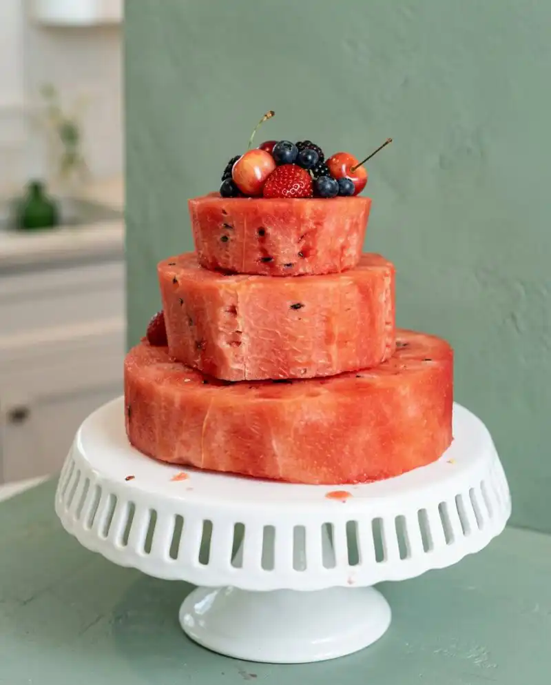 A three tier Fresh Watermelon Layer Cake decorated with mixed berries and mint leaves on a white stand.
