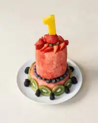 A beautiful tiered Baby's First Watermelon Yogurt Cake covered in creamy frosting and fresh berries.