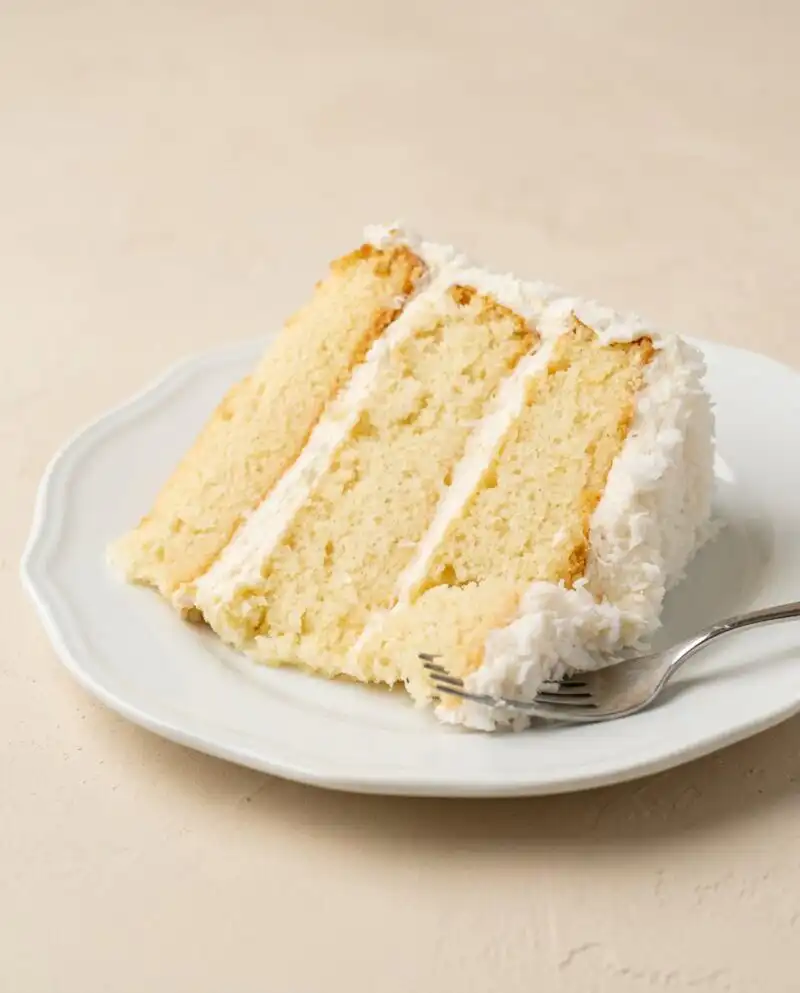 A beautiful Three-Layer Coconut Cream Cake coated in sweet shredded coconut on a white cake stand.
