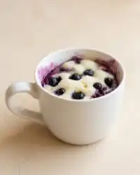 A warm and fluffy easy blueberry mug cake served in a dark ceramic mug on a wooden table.