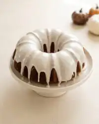 A glazed pumpkin spice bundt cake sitting on a white cake stand in a bright kitchen setting